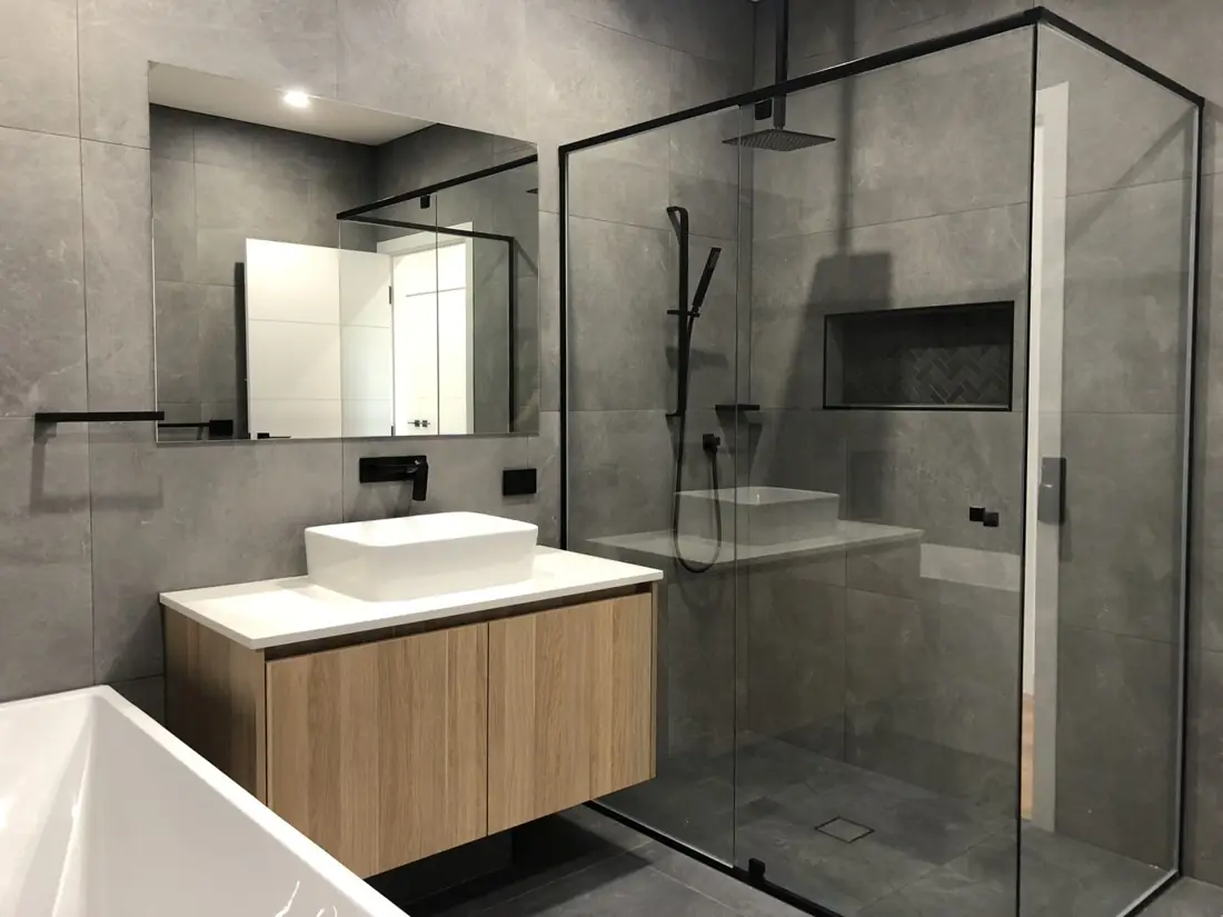 Grey tiled bathroom with black-framed shower screen, timber vanity and bath